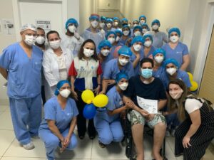 Colaboradores do ISAC no Hospital Estadual cercam o paciente Francisco Leandro para se despedirem dele durante a alta médica. Ele está à direita, sentado em uma cadeira de rodas, acompanhado da mulher que aparece agachada do lado esquerdo dele. Alguns colaboradores seguram balão.