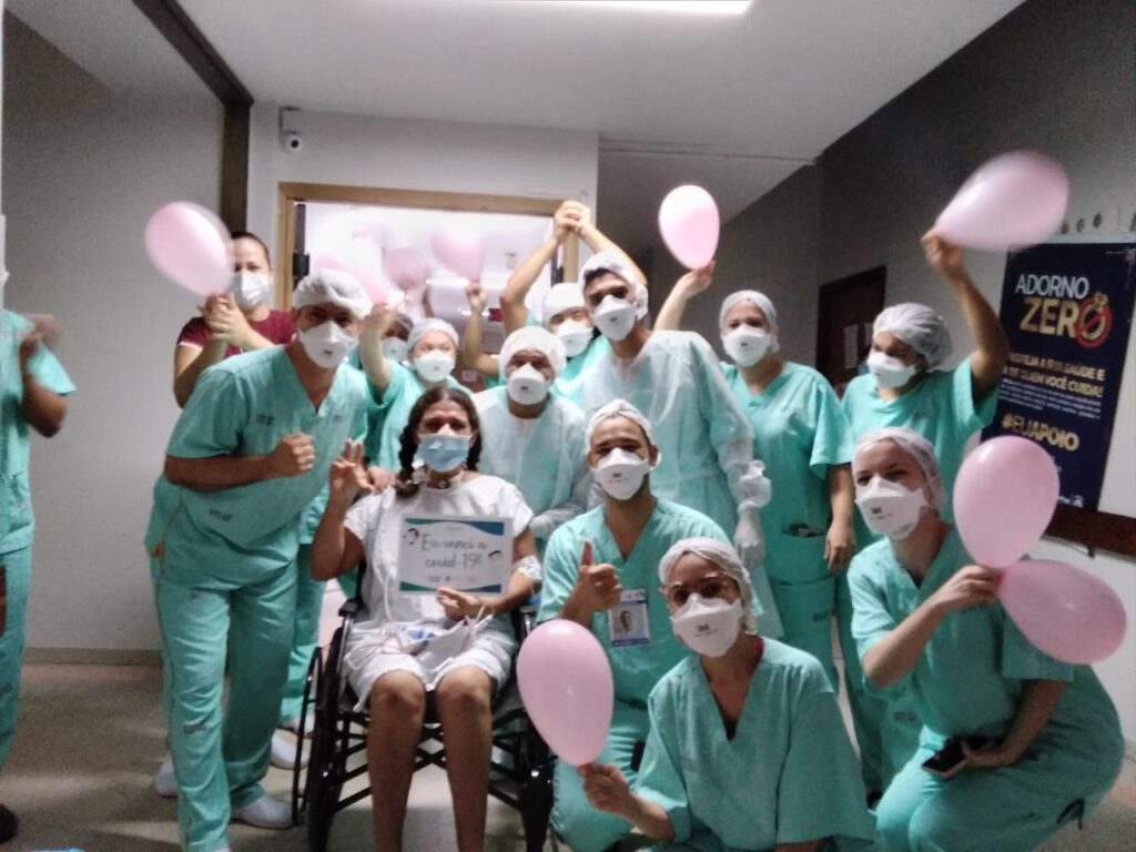 Várias colaboradores estão em volta de uma paciente. Ela está sentada em uma cadeira de rodas e segura um cartaz onde está escrito que venceu a covid-19. Eles seguram balões cor de rosa.
