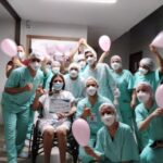 Várias colaboradores estão em volta de uma paciente. Ela está sentada em uma cadeira de rodas e segura um cartaz onde está escrito que venceu a covid-19. Eles seguram balões cor de rosa.