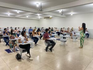 Imagem de um auditório grande. Do lado direito estão diversos profissionais sentados em cadeiras dispostas com uma certa distância entre elas. Todas as pessoas usam máscara. Na frente deles, do lado direito da foto, aparece a ministrante do curso. Ela está de perfil. Tem cabelos longos e pretos e veste um macacão estampado verde e branco.