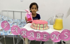 Foto de uma menina atrás de uma mesa com um bolo, uma jarra de suco e uma faixa onde está escrito "parabéns". Ela aparece do peito para cima, segura um presente, tem pele de cor negra e cabelos pretos. Atrás dela há uma cama de hospital.