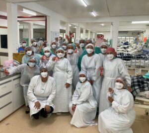 17 colaboradoras do ISAC posam de frente para a câmera. Todas usam uniforme, máscara facial e touca de cabelo. Elas estão ao lado de um balcão na UTI Covid-19 do Hospital Regional de Gurupi. Algumas em pé, outras agachadas. Algumas seguram presentes recebidos pela ocasião do Dia da Mulher. Atrás delas há um enorme salão com leitos espalhados.