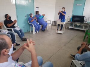 Em destaque, no fundo da sala, uma mulher está em pé. Ela une as duas mãos. O movimento é copiado pelos demais participantes do curso que a rodeiam. Eles estão sentados em cadeiras brancas.