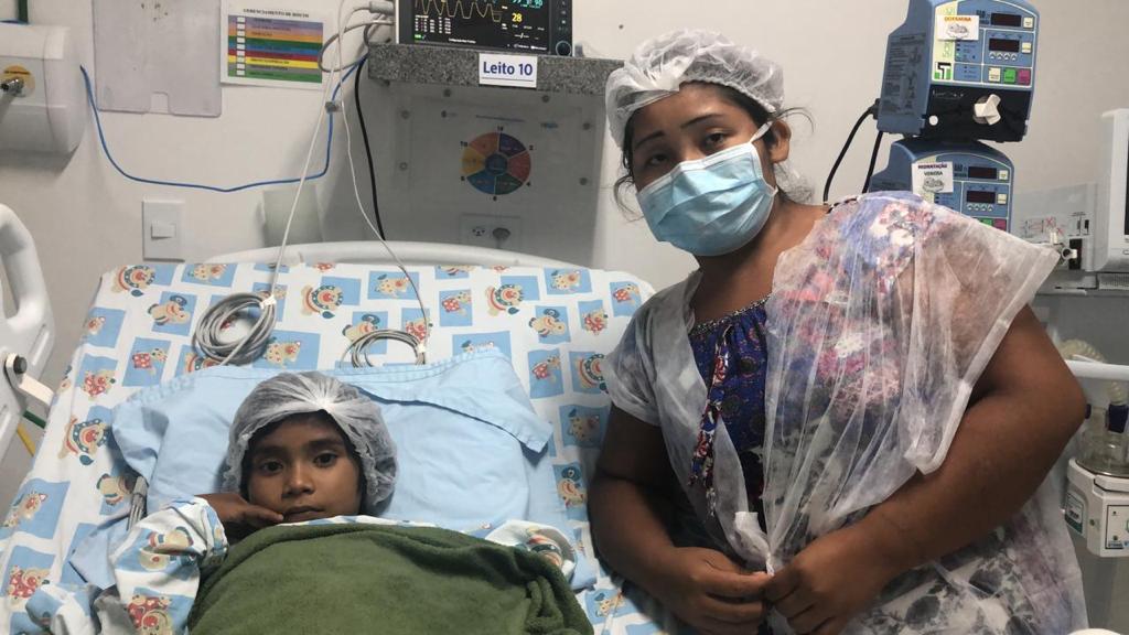 Foto da paciente com a mãe. A menina está deitada e embrulhada até o pescoço. Ela olha sério pra câmera e usa touca na cabeça. A mãe está debruçada no leito, do lado direito da filha. Ela usa touca, máscara e avental. Atrás delas há vários equipamentos hospitalares, como monitores e régua de gás.