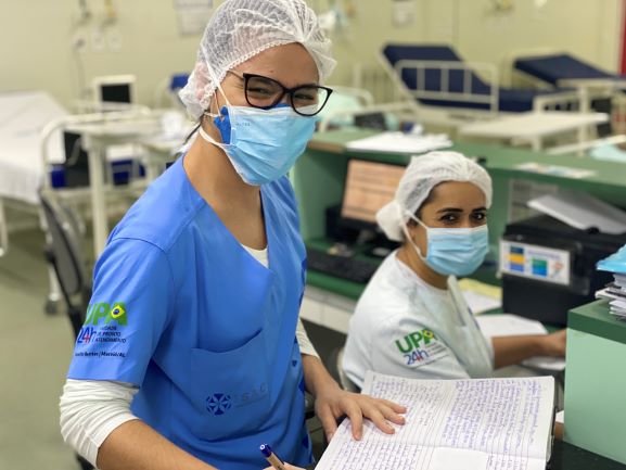 Duas mulheres em um posto de enfermagem. Elas estão uniformizadas, usam máscara facial e sorriem para a câmera.