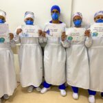 Cinco profissionais da saúde, uniformizados com máscara, touca e avental. Eles seguram placas com mensagens sobre higiene das mãos. Estão lado a lado, de frente para a câmera, no meio de um corredor.