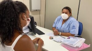 Em primeiro plano há uma mulher de cor de pele negra. Ela está de costa para a câmera e aparece de perfil, com as mãos sobre uma mesa. Na frente dela há uma mulher, que usa máscara facial branca e uniforme azul. Na mesa há um computador e muitos papéis espalhados.