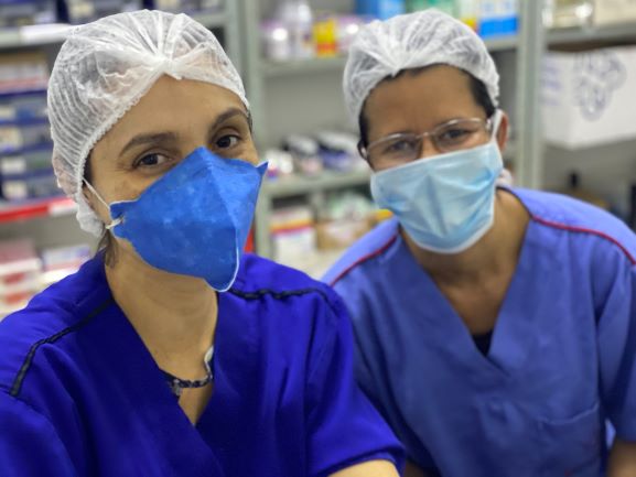 Duas mulheres sentadas na sala da farmácia do hospital. Elas usam uniforme azul, máscara facial e touca. Ambas sorriem para a câmera.