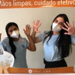 Duas mulheres posam de frente para a câmera com as mãos levantadas. Na frente delas há uma moldura laranja onde está escrito: "Mãos limpas, cuidado efetivo".