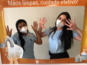 Duas mulheres posam de frente para a câmera com as mãos levantadas. Na frente delas há uma moldura laranja onde está escrito: "Mãos limpas, cuidado efetivo".