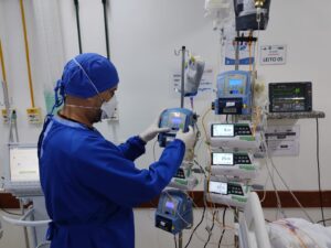 Um enfermeiro de perfil. Ele usa luvas, avental, máscara e touca. Manuseia um equipamento.