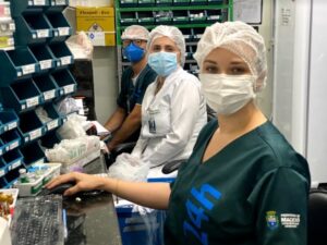 Três pessoas sentadas de lado olham para a câmera.Elas estão dentro da farmácia da UPA.