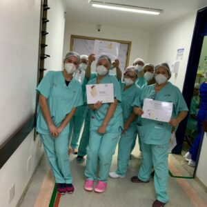 Imagem de um grupo de colaboradores em um corredor. Todos vestem uniforme verde da assistência e usam touca e máscara.