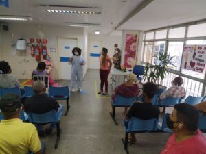 Foto da recepção do Multicentro Carlos Gomes. Em primeiro plano há vários pacientes sentados e, ao fundo, duas profissionais da unidade em pé.