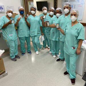 Oito profissionais estão lado a lado formando uma meia lua. Todos usam uniforme verde claro, touca, máscara facial e seguram uma caneca nas mãos.