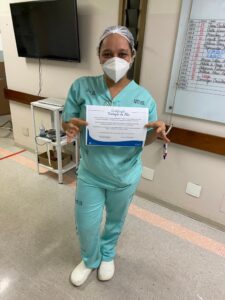 Mulher com fardamento hospitalar posa para foto segurando o certificado do destaque do mês.
