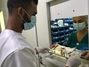 Homem aparece de perfil, usando máscara e luvas, recebendo uma bandeja com materiais hospitalares por uma janela interna do hospital. Do outro lado, uma mulher de máscara e farda verde, estrega a bandeja ao homem.