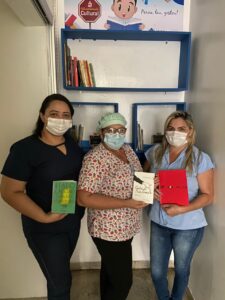 Três mulheres posam para foto. Cada uma segura um livro nas mãos. Todas usam máscara e sorriem oara a câmera.