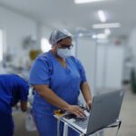 mulher com touca, máscara e fardamento hospitalar na cor azul maneja aparelhagem hospitalar. Ao fundo, com imagem desfocada, mostra uma ambiente de hospital.