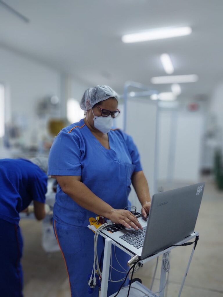 mulher com touca, máscara e fardamento hospitalar na cor azul maneja aparelhagem hospitalar. Ao fundo, com imagem desfocada, mostra uma ambiente de hospital.