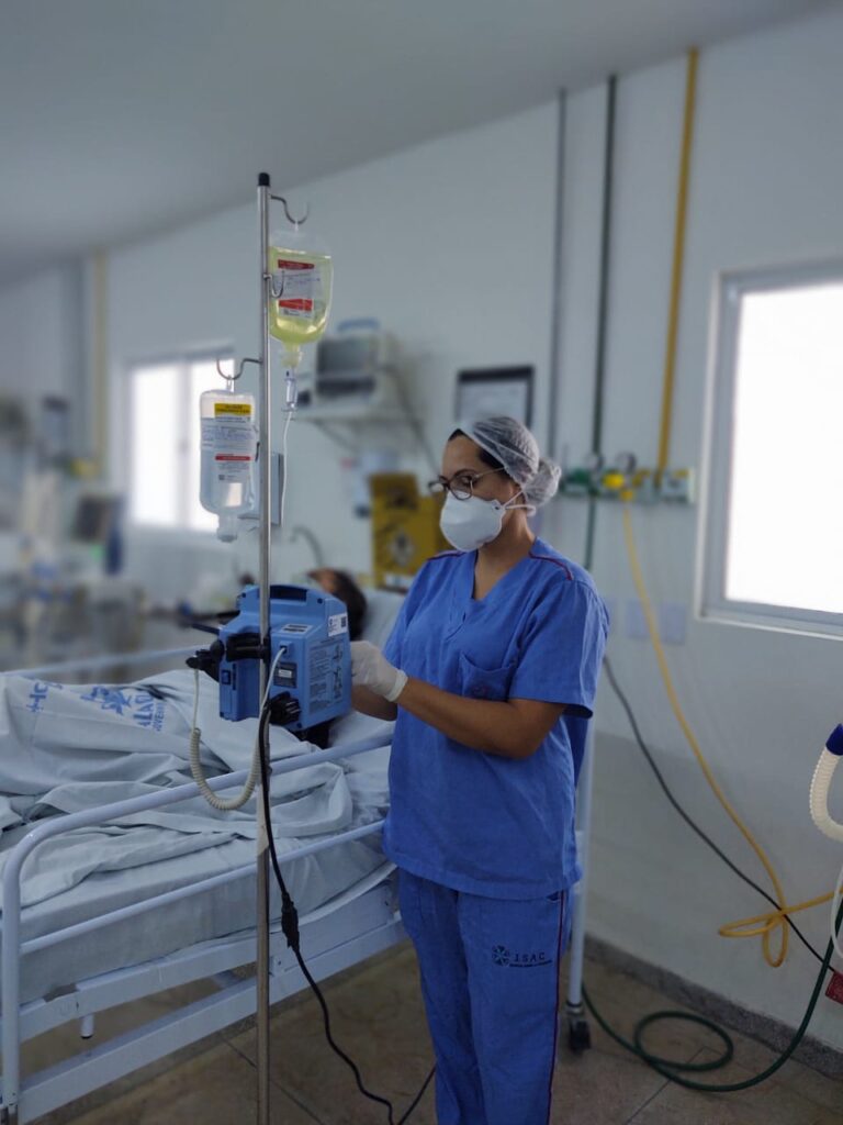 Uma mulher está de pé ao lado de um leito hospitalar. Ela usa fardamento azul, está de máscara, touca e luvas. A mesma maneja um aparelho hospitalar.