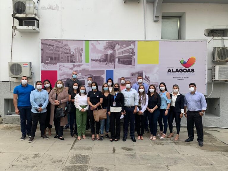 Várias pessoas, uma ao lado da outra, posam para câmera. Todas usam máscara. Ao fundo, um painel com fotos de unidades de saúde e a marca da Prefeitura do Governo do Estado de Alagoas.
