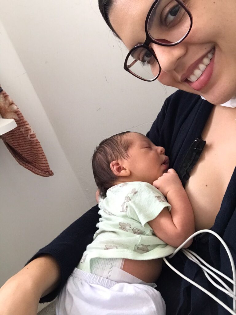 Bebê está dormindo no colo da mãe. Ela está sentada, usa óculos de grau e sorri para câmera.