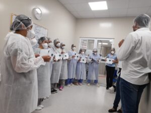 Algumas pessoas aparecem de pé em um corredor hospitalar, uma ao lado da outra formando um semicírculo. Elas estão de avental, touca, máscara e seguram uma caixa para descarte de materiais perfurocortantes.