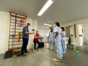 Mulher realiza exercício físico enquanto quatro pessoas com fardamento hospitalar observam a atividade.