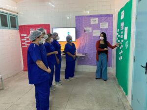 Uma mulher aparece ao lado de um mural verde. Ao lado dela, algumas pessoas com fardamento hospitalar azul, máscara e touca observam o que está sendo apresentado no mural.