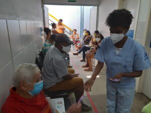 Mulher com farda hospitalar azul claro e máscara está de pé e entrega folhetos informativos. Ao redor dela, várias pessoas estão sentadas em cadeiras.