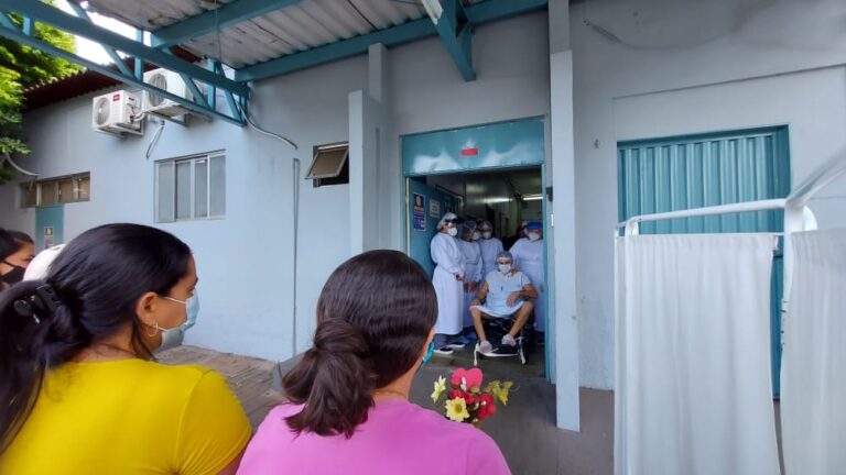 Em primeiro plano, duas mulheres aparecem de costas. Uma delas segura flores. ao fundo, um homem sentado em uma cadeira de rodas olha emocionado para as mulheres.