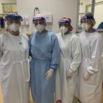 Quatro mulheres estão de pé e posam para foto. Todas estão com avental hospitalar, touca, máscara, protetor facial e luvas. Ao fundo, aparece leitos hospitalares de UTI.