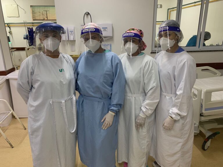 Quatro mulheres estão de pé e posam para foto. Todas estão com avental hospitalar, touca, máscara, protetor facial e luvas. Ao fundo, aparece leitos hospitalares de UTI.