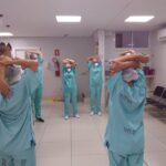 Trabalhadores da saúde fazem exercício laboral dentro de ambiente hospitalar. Todos estão com uniforme verde e, usam touca e máscara.