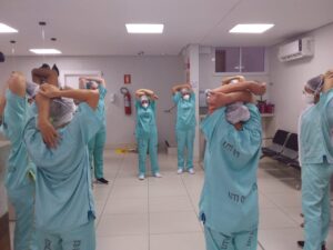 Trabalhadores da saúde fazem exercício laboral dentro de ambiente hospitalar. Todos estão com uniforme verde e, usam touca e máscara.