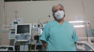 Uma mulher com uniforme verde, touca e máscara hospitalar posa para câmera com os braços para trás. Ao fundo, aparecem aparelhos e leitos hospitalares .