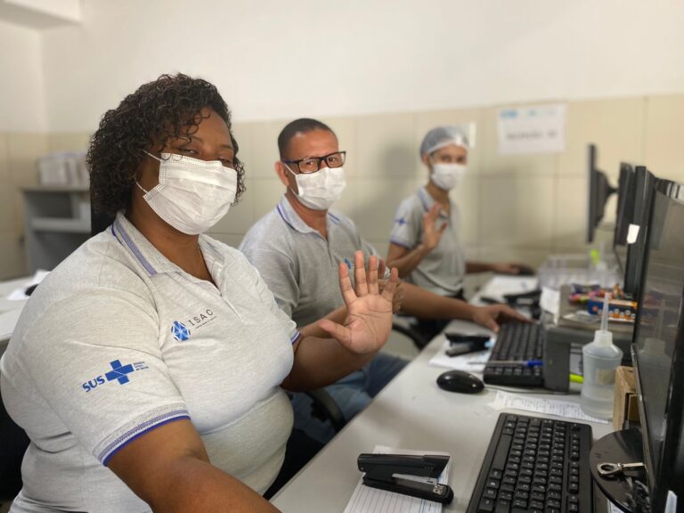 Três pessoas sentadas, uma ao lado da outra, em frente a computadores. Todos estão de máscara e camisa polo cinza com as logomarcas do SUS e do ISAC