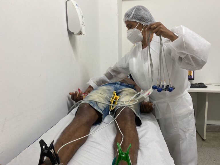 Um homem está deitado no leito com vários eletrodos pelo corpo. De pé, uma profissional de saúde com touca, luva e máscara realiza o manejo do paciente.