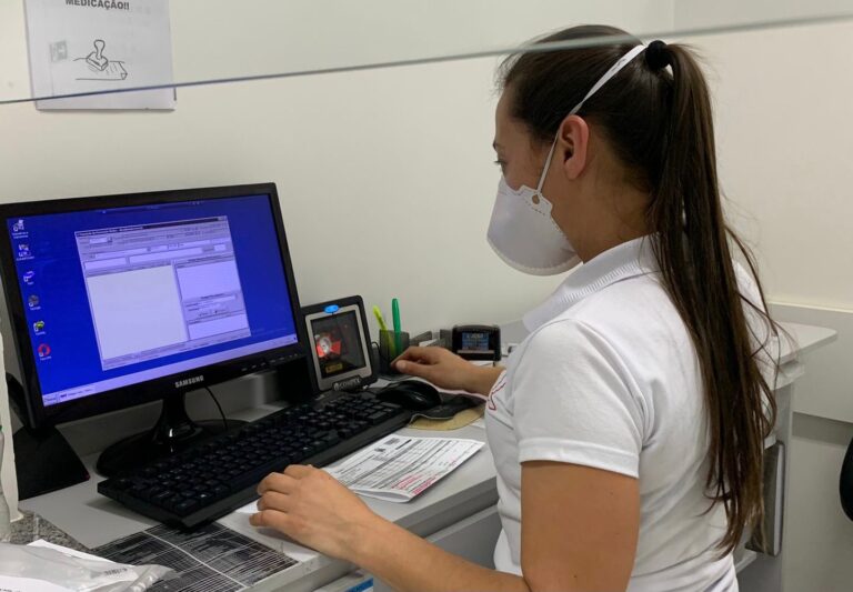 Mulher aparece sentada em frente a uma tela de computador e de costas para câmera. Ela usa máscara.