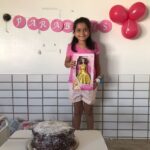 Uma menina está de pé em frente a uma mesa com um bolo de aniversário. Ela segura uma boneca e na parede tem balões rosas com uma faixa escrita feliz aniversário.