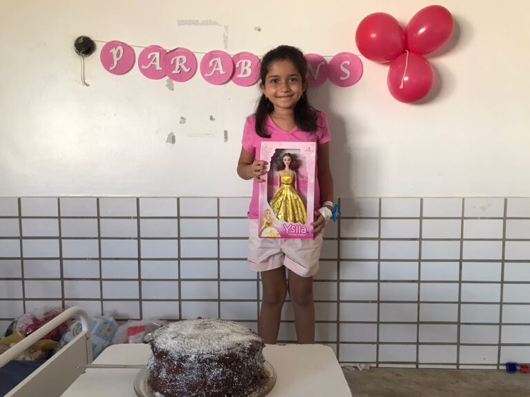 Uma menina está de pé em frente a uma mesa com um bolo de aniversário. Ela segura uma boneca e na parede tem balões rosas com uma faixa escrita feliz aniversário.