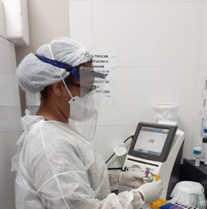 Uma mulher com avental hospitalar, máscara, luvas e touca está manipulando um tubo de ensaio.
