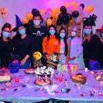 Foto da comemoração dos aniversariantes do mês na UPA Cidade Nova. Há oito pessoas atrás de uma mesa cheia de doces e bolos. A decoração é laranja e roxa, com símbolos do Dia das Bruxas, como chapéu de bruxa e abóbora com rosto.