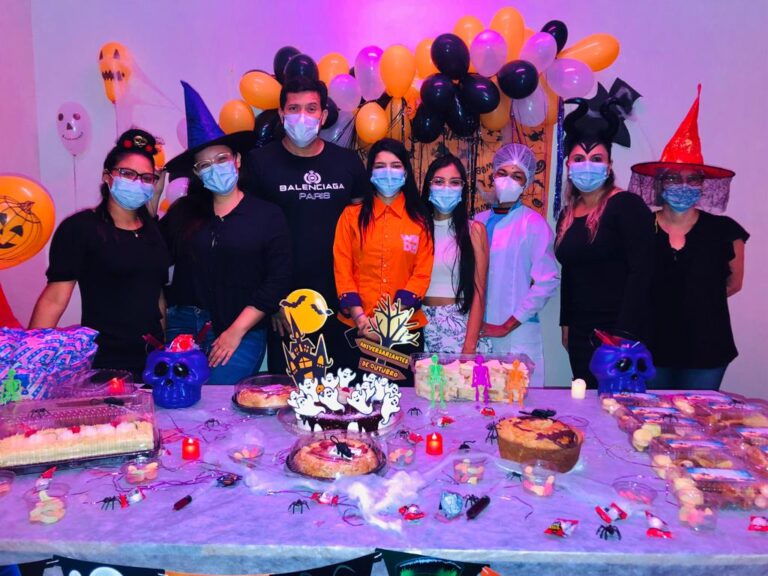 Foto da comemoração dos aniversariantes do mês na UPA Cidade Nova. Há oito pessoas atrás de uma mesa cheia de doces e bolos. A decoração é laranja e roxa, com símbolos do Dia das Bruxas, como chapéu de bruxa e abóbora com rosto.