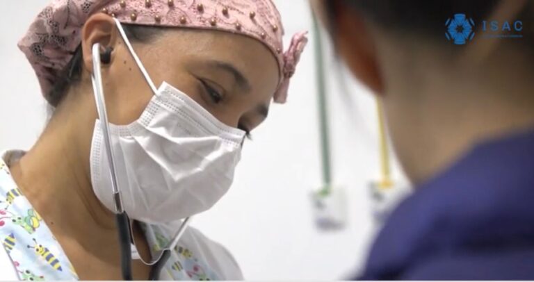 Uma mulher de máscara de touca usa estetoscópio. De costas, aparece a nuca da paciente.