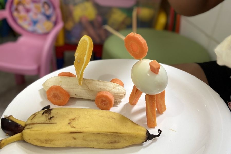Imagem mostra um prato com uma banana com casca e outras duas bananas enfeitadas no formato de carro e cachorro.
