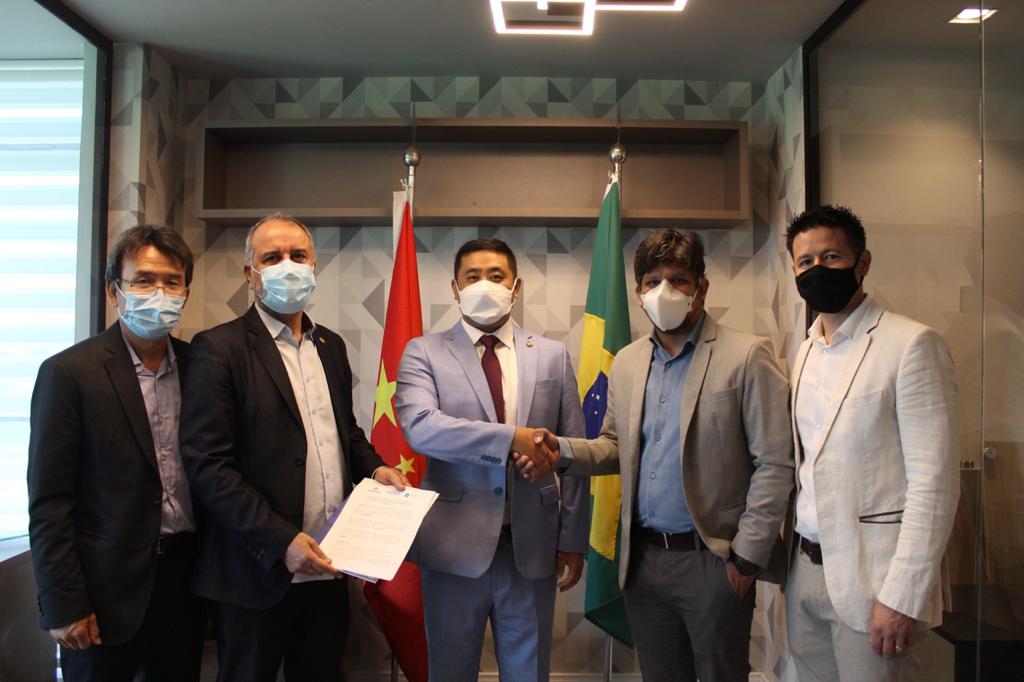 A imagem mostra cinco homens posando para câmera, um ao lado do outro. Todos usam terno e máscara. Os dois do centro apertam as mãos. Ao fundo está uma bandeira do Brasil e outra da China.
