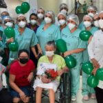 Diversos profissionais, devidamente paramentados e com máscara, estão com balões verdes ao redor da cadeira de rodas da paciente.