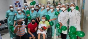 Diversos profissionais, devidamente paramentados e com máscara, estão com balões verdes ao redor da cadeira de rodas da paciente.